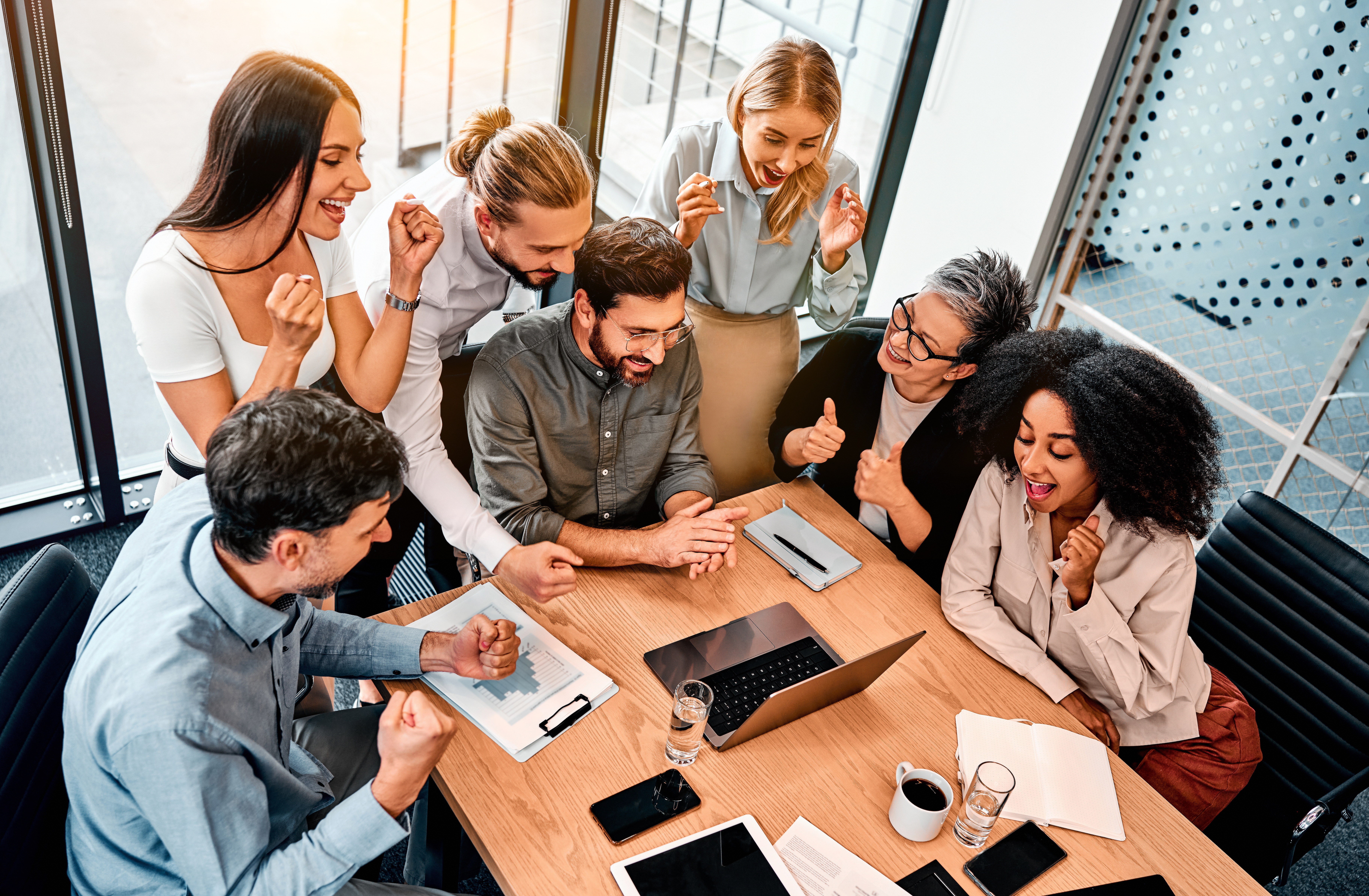 Cheering for colleague. View from above on business team gathering around caucasian man sitting by wooden table in front of laptop. Supportive people believing in success of newly hired employee.