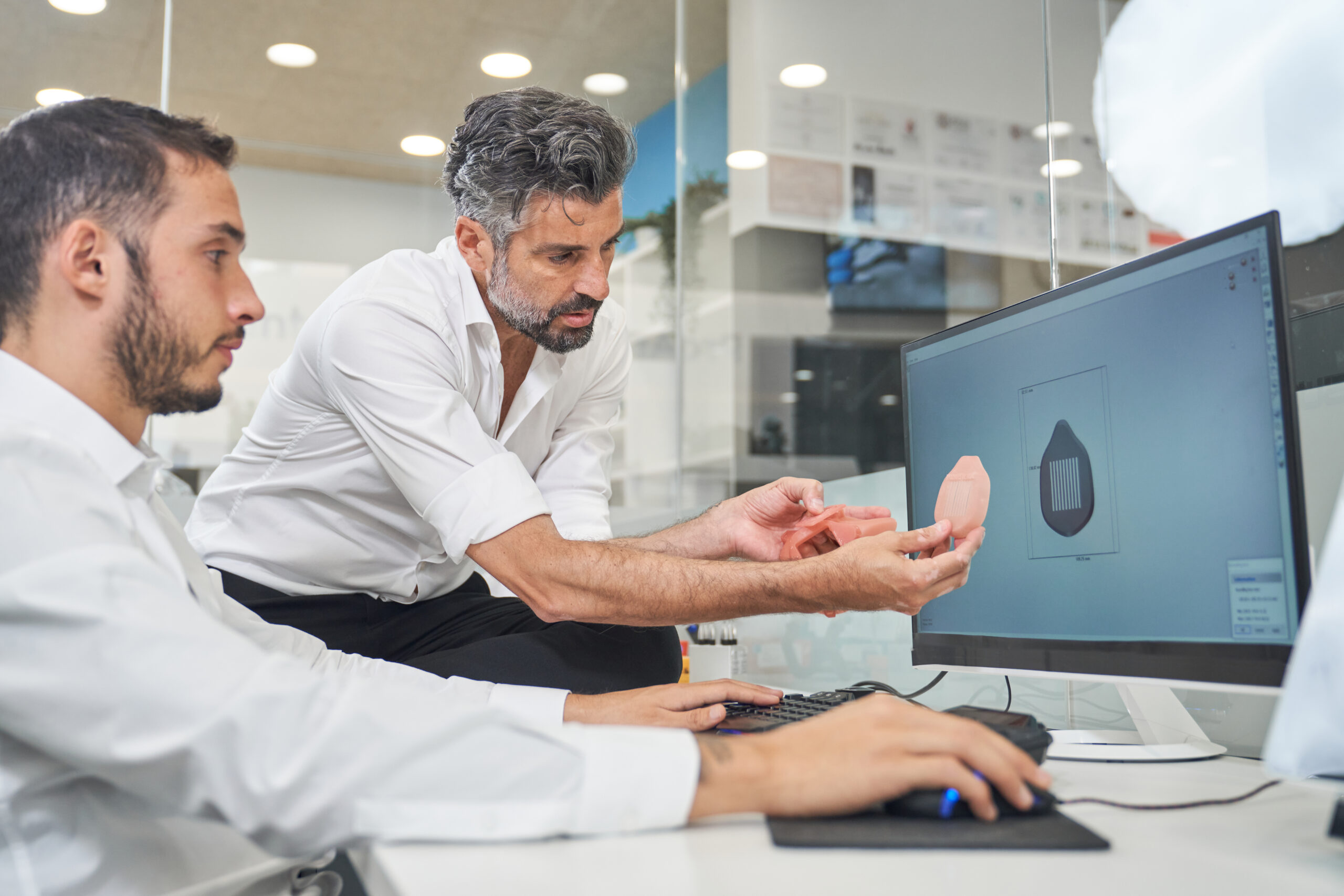 Concentrated bearded male researchers examining printed face mask model while working on computer with 3D illustration of respirator in contemporary office
