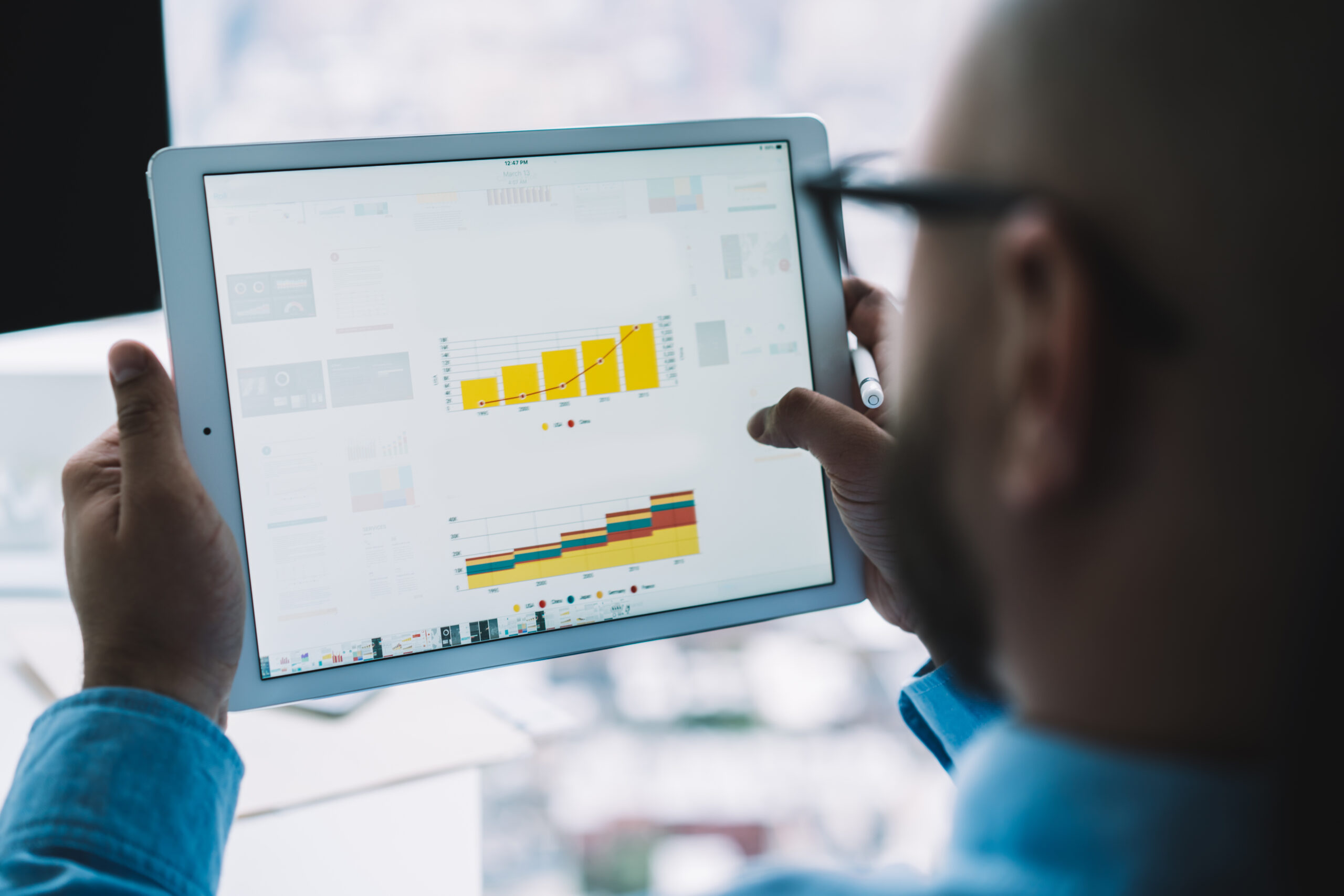 Crop blurred man in glasses holding modern tablet and exploring colorful graph templates while working with statistics in light office