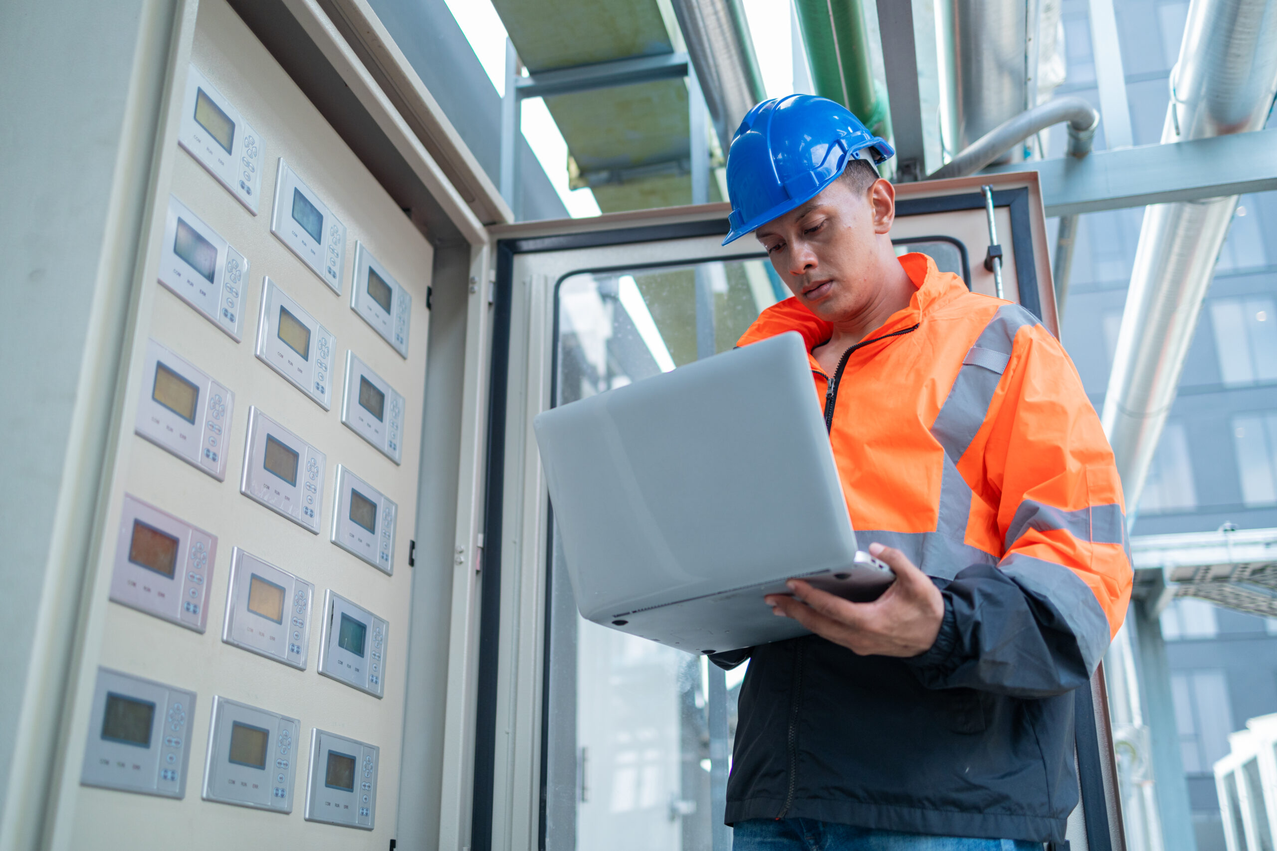A technician in an orange high-visibility jacket and blue hard hat operates a control panel while referencing a laptop at an industrial site.