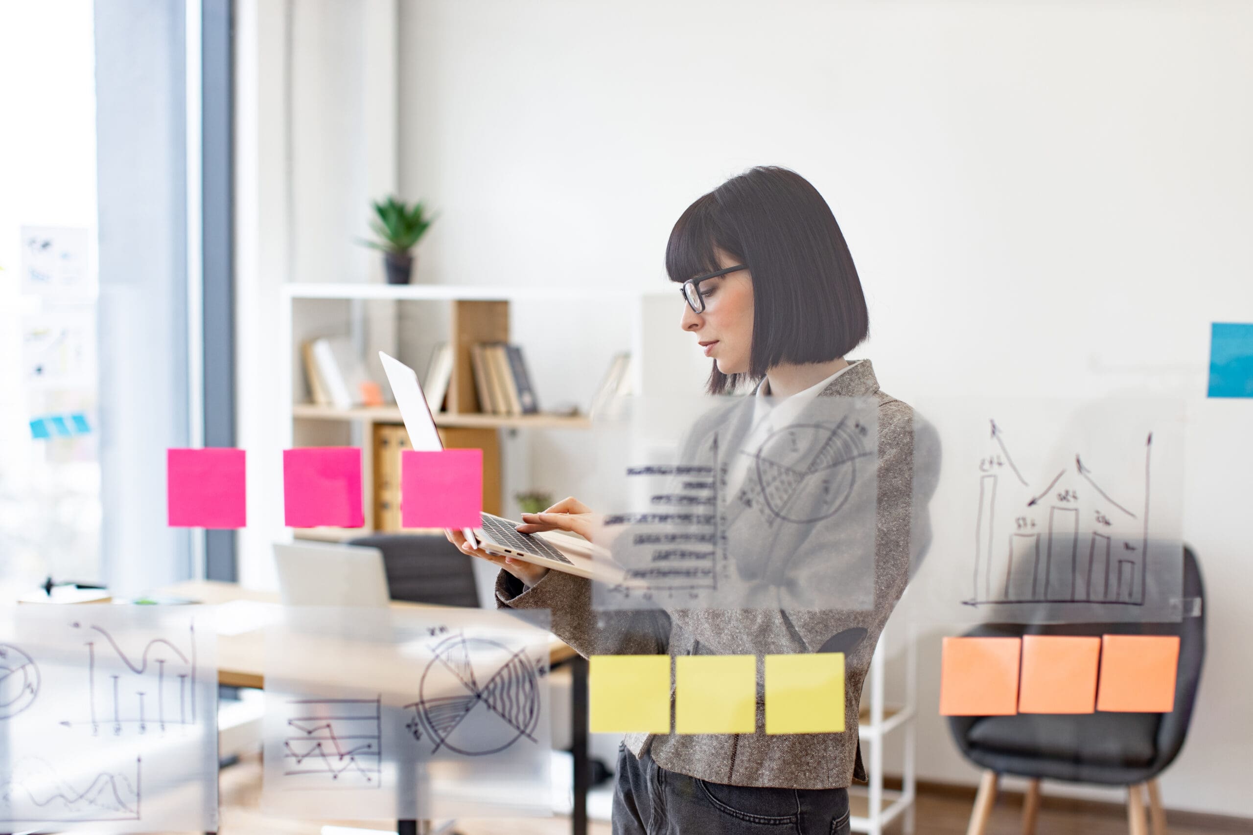 Pensive businesswoman using digital laptop while analyzing growth of charts on glass board at modern office. Concept of business strategy and digital marketing.
